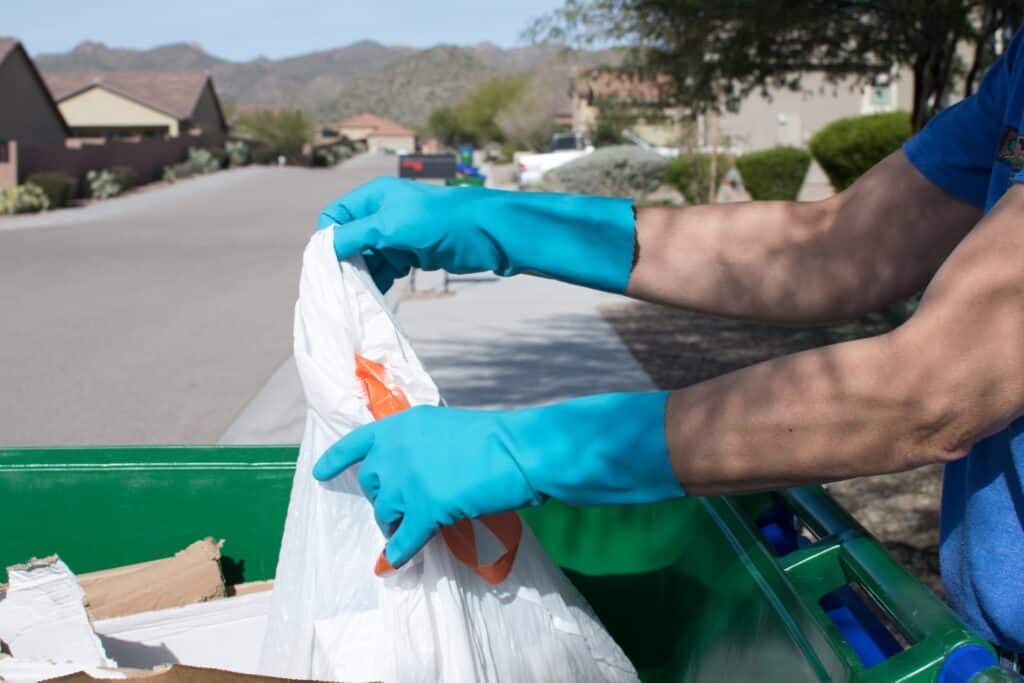 Person wearing blue gloves disposing of junk into a green dumpster.