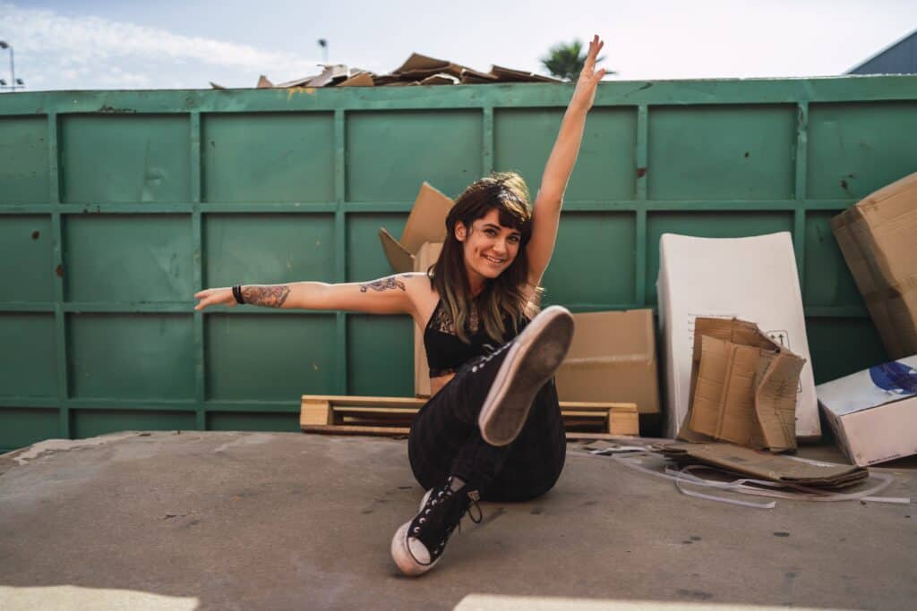 A woman smiling and sitting in front of a large green dumpster filled with cardboard boxes and debris, showcasing junk remova.