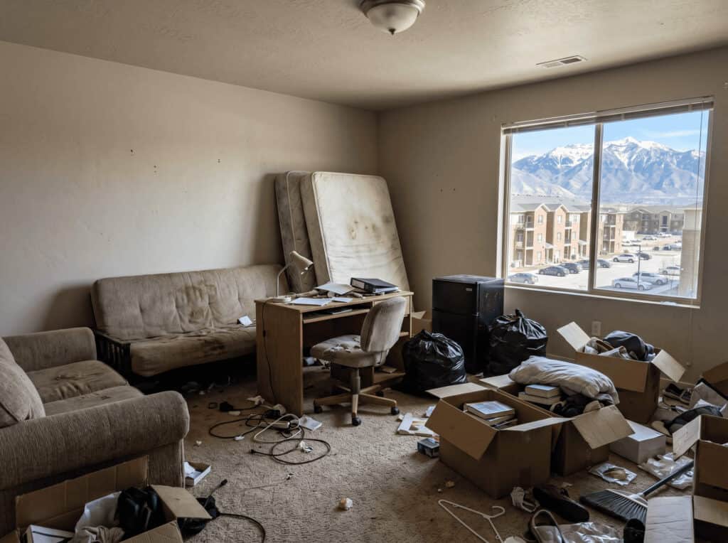 Interior of a cluttered living room with furniture, boxes, and trash, overlooking snow-capped mountains through a large windo.